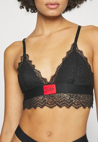 Zwarte kanten bralette met een diepe halslijn, voorzien van golvende randen, doorzichtige textuur en een prominente rode band met het "HUGO" label.