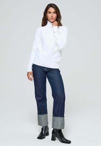 Un pull col roulé blanc, ajusté, associé à un jean bleu foncé retroussé. Le mannequin porte des bottes à talons noires, se tenant devant un fond neutre.