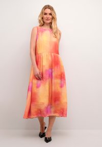 Satin ærmeløs kjole i orange, pink og gul ombre mønster. A-line silhuet med samlet talje. Sorte spidse stiletter.