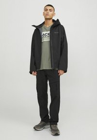 Schwarze wasserdichte Jacke mit Kapuze, Reißverschluss vorne und Seitentaschen, getragen über einem olivgrünen T-Shirt mit Grafik und schwarzen Cargo-Hosen.