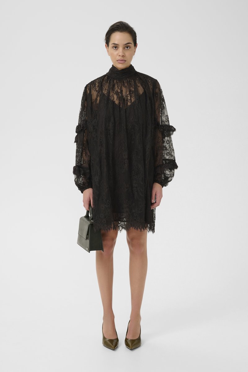 Black lace dress with high collar, long sleeves, and ruffled accents. Worn with olive green heels and a small dark green handbag.