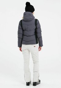 Daunenjacke in Dunkelgrau mit schwarzen Akzenten, ausgestattet mit einer Kapuze und quilteter Textur. Getragen mit weißen Hosen und schwarzen Skischuhen.