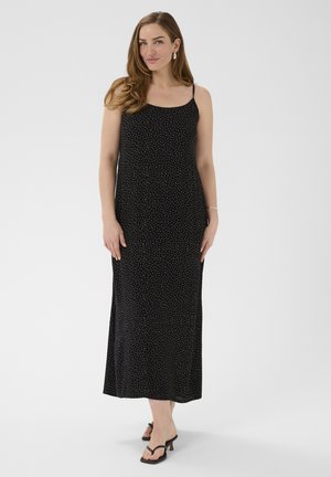 Femme portant une robe maxi noire sans manches à petits pois blancs, debout en posture détendue avec des sandales noires à talons sur un fond blanc.