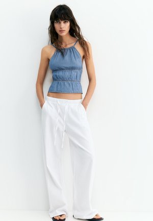 Femme aux longs cheveux bruns portant un top bleu sans manches et un pantalon blanc ample, debout les mains dans les poches contre un mur blanc.