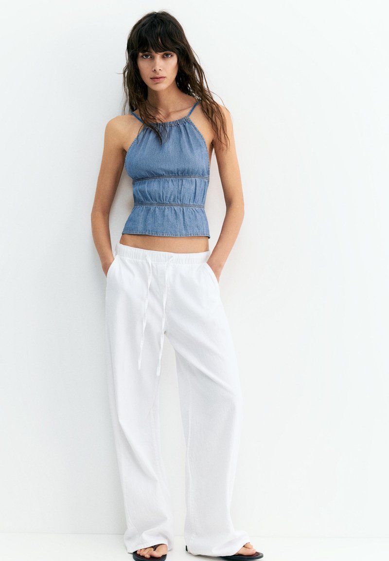 Femme aux longs cheveux bruns portant un top bleu sans manches et un pantalon blanc ample, debout les mains dans les poches contre un mur blanc.