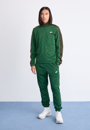 Costum sport verde compus dintr-o jachetă cu fermoar și guler înalt, cu un logo alb, și pantaloni asortați cu manșete elastice, completat de adidași albi.
