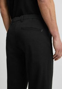 Schwarze Hose mit geradem Schnitt, ausgestattet mit einer Reißverschlusstasche auf der Rückseite und einer glatten, matten Textur. Der Bund ist schlicht und ohne sichtbare Verschlüsse.