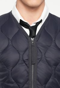 Ναυτικό γουνάκι με φερμουάρ, με λείο, γυαλιστ�ερό υφή και διαμαντένιο μοτίβο, φοριέται πάνω από ένα λευκό πουκάμισο και μαύρη γραβάτα.