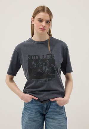 Junge Frau mit langen blonden Haaren, die ein dunkelgraues T-Shirt mit Grafik und hellblaue Jeans trägt, Hände in den Taschen, neutraler Hintergrund.