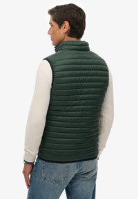 Groene puffer-vest met horizontaal gestikt patroon, hoge kraag en zwarte rand. Gedragen over een crèmekleurig textuurachtige lange mouwen shirt, gecombineerd met blauwe spijkerbroek.