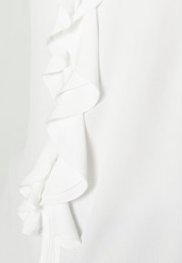 Biały materiał z warstwowymi falbankami wzdłuż pionowej krawędzi, ukazujący teksturę i miękkie fałdy.