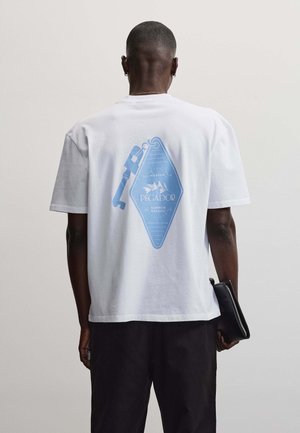 Man draagt wit t-shirt met blauw sleutel- en labelontwerp op de rug, zwarte broek, houdt een zwarte etui met rits vast, met de rug naar de camera.