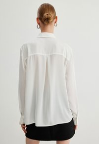Blusa blanca de manga larga, fabricada en una tela ligera, con un suave brillo, detalle de pliegue en la parte posterior y un cuello clásico, combinada con una falda negra texturizada.