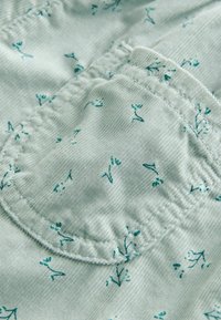 Hellgrüner strukturierter Stoff mit kleinen blauen Blumenmustern. Merkmale sind eine Tasche mit sichtbarenNähten und elastischer Raffung.