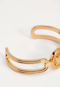 Brazalete de oro con diseño abierto de doble banda y medallón circular grabado con la cabeza de un león sobre fondo blanco.