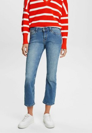 Personne portant un jean court bleu, des baskets blanches et un pull à manches longues rayé rouge et blanc, debout devant un fond uni.