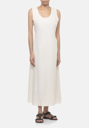 Donna che indossa un vestito bianco senza maniche fino alla caviglia abbinato a sandali neri piatti su uno sfondo chiaro uniforme.