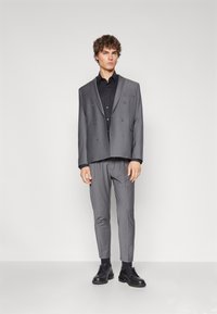 Grå dobbeltradet blazer med matt finish, kombinert med slim-fit grå bukser og svarte snørestøvler. Svart skjorte under.