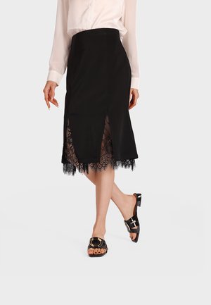 Donna che indossa una gonna midi nera con bordo in pizzo e spacchi frontali abbinata a una camicetta bianca e sandali neri con tacco.