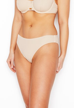 Donna che indossa un bikini beige testurizzato con slip e reggiseno a ferretto abbinati, in piedi davanti a uno sfondo bianco.