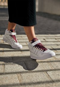 Höga sneakers i vit läder med vinröda snören och detaljer. Har ett kardborreband och guld detaljer mot en kullerstensbakgrund.