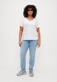 Camiseta de algodón blanca con cuello en V y logo rojo, combinada con jeans de color azul claro y zapatillas grises. Cabello rizado y texturizado y gafas de sol.
