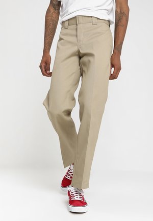 Personne portant un pantalon beige, un t-shirt blanc et des baskets rouges marchant devant un fond clair uni.