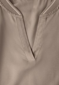 Vêtement à col en V en tissu beige doux, présentant une finition de couture propre et une texture lisse. Design simple sans motifs ni matériel visible.