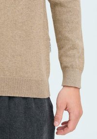 Beige, hosszú ujjú pulóver bordás mandzsetta és alja. Puha textúra, oldalsó címke látható. Sötétszürke nadrággal párosítva.