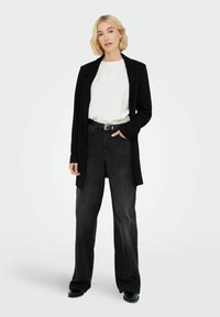 Kvinna med kort blont hår som bär en vit skjorta, svart kavaj, vida svarta jeans och svarta stövlar, stående med ena handen i fickan.