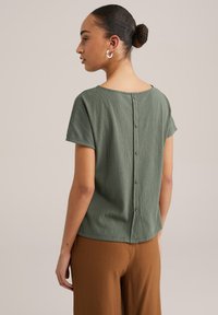 Zaļa īsām piedurknēm topa, kas izgatavots no mīksta auduma ar apaļu kakla izgriezumu un pogām aizmugurē. Saskaņots ar brūnām teksturētām biksēm.