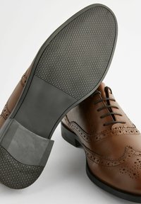 Chaussures de ville en cuir marron avec des détails en brogue, une tige lisse et une semelle en caoutchouc texturée avec un talon superposé.