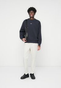 Sweatshirt azul-marinho com logotipo branco, calças creme e sapatos pretos. O modelo segura uma pequena bolsa preta e está de pé contra um fundo branco.