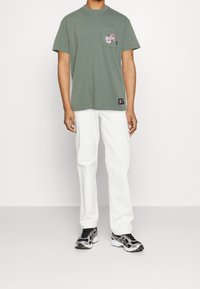 Green cotton t-shirt with a chest pocket featuring a graphic design, paired with white corduroy pants and black athletic sneakers.