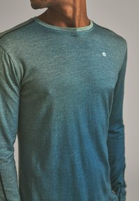 Long-sleeve shirt in gradient teal fabric, featuring a textured pattern, round neckline, and subtle logo on the chest. Accented with black shoulder seams.