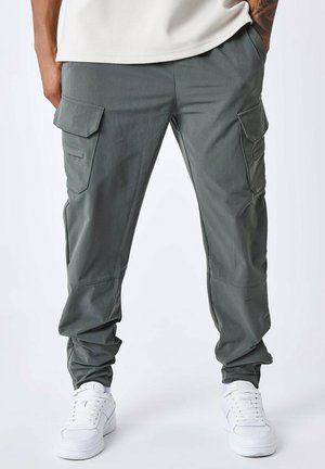 Pantalones cargo en verde oliva con múltiples bolsillos laterales, puños elásticos y un corte relajado, combinados con zapatillas blancas. Textura de tela suave.