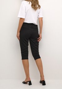 Černé zkrácené kalhoty s pružným pasem, se dvěma zadními kapsami a hladkým povrchem. Nošeny s bílým tričkem a černými botami na pevné podrážce s blokovým podpatkem.