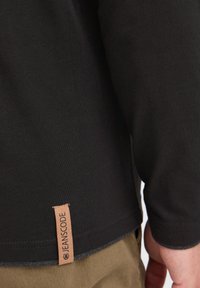 Zwarte langslavende shirt van textuurstof met een bruine leren label met de tekst "JEANSCODE" aan de onderrand.