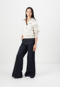 Beige zip-up sweatshirt with wide collar, paired with dark blue wide-leg jeans. Plain texture with visible stitching and white sneakers.