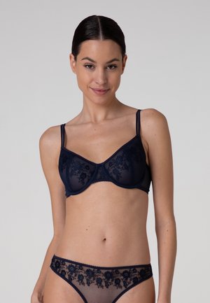 Donna con capelli scuri che indossa reggiseno e slip in pizzo blu navy coordinati, sorridendo leggermente, su sfondo chiaro e uniforme.
