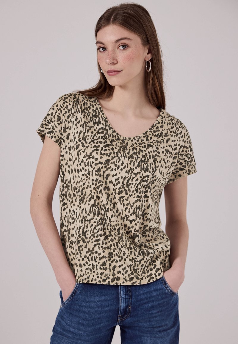 Beige Oberteil mit kurzen Ärmeln, das ein schwarzes und olivgrünes Leopardenmuster, einen runden Ausschnitt und einen lockeren Schnitt aufweist. Kombiniert mit blauen Jeans.