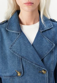 Denim trenchcoat i mellomblå, med dobbeltradet design, store brune knapper og markante krager. Teksturlignende stoff med synlig søm.
