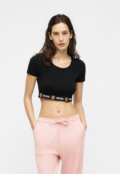 Γυναίκα που φοράει μαύρο κοντό μπλουζάκι με λωρίδα με λογότυπο αρκούδας και ανοιχτό ροζ παντελόνι με κορδόνι, στέκεται με τα χέρια στις τσέπες.