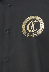 Camisa negra abotonada con un logo circular dorado que presenta una "C" estilizada y el texto "JUST CAVALLI" en el interior, con detalles texturizados de diseño de serpiente.