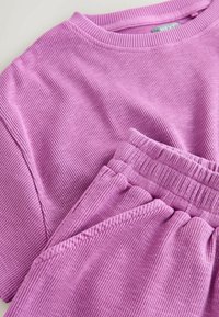 Měkké fialové tričko s krátkými rukávy a žebrovaným úpletem a k němu odpovídající kalhoty s elastickým pasem, položeny na rovné ploše, zobrazující texturu látky a detaily švů.