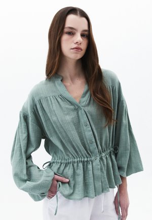 Mintgroene blouse met knoopsluiting, met een gegatherde taille, lange mouwen en een gestructureerde stof. Heeft een V-hals en strikjes aan de zijkanten.