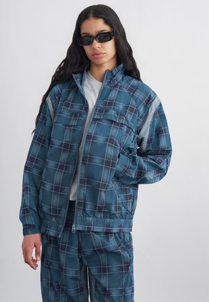 Jeune femme aux longs cheveux foncés portant des lunettes de soleil noires et une veste et un pantalon à carreaux bleus, debout devant un fond uni clair.