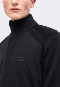 Νεαρός άνδρας που φοράει μαύρο μπουφάν με ψηλό γιακά και υφασμάτινες πλάκες στους ώμους, με ανάγλυφα λογότυπα Porsche BOSS στο στήθος.