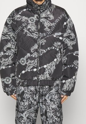 Ensemble veste matelassée noire et pantalon avec motifs floraux baroques gris et chaînes, comprenant le texte « Versace Jeans Couture » dans différentes orientations.