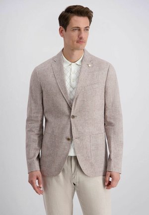 Man draagt een lichtbeige getextureerd colbert over een wit poloshirt met patroon en beige broek, staand tegen een effen achtergrond.